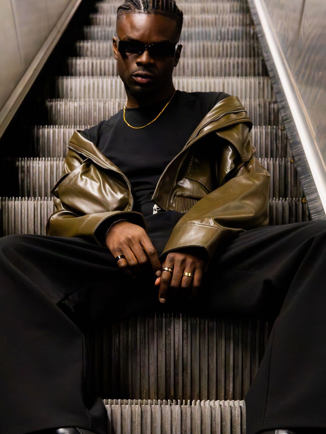 Graël assis dans un escalier mécanique, veste de cuir brun et lunettes noires